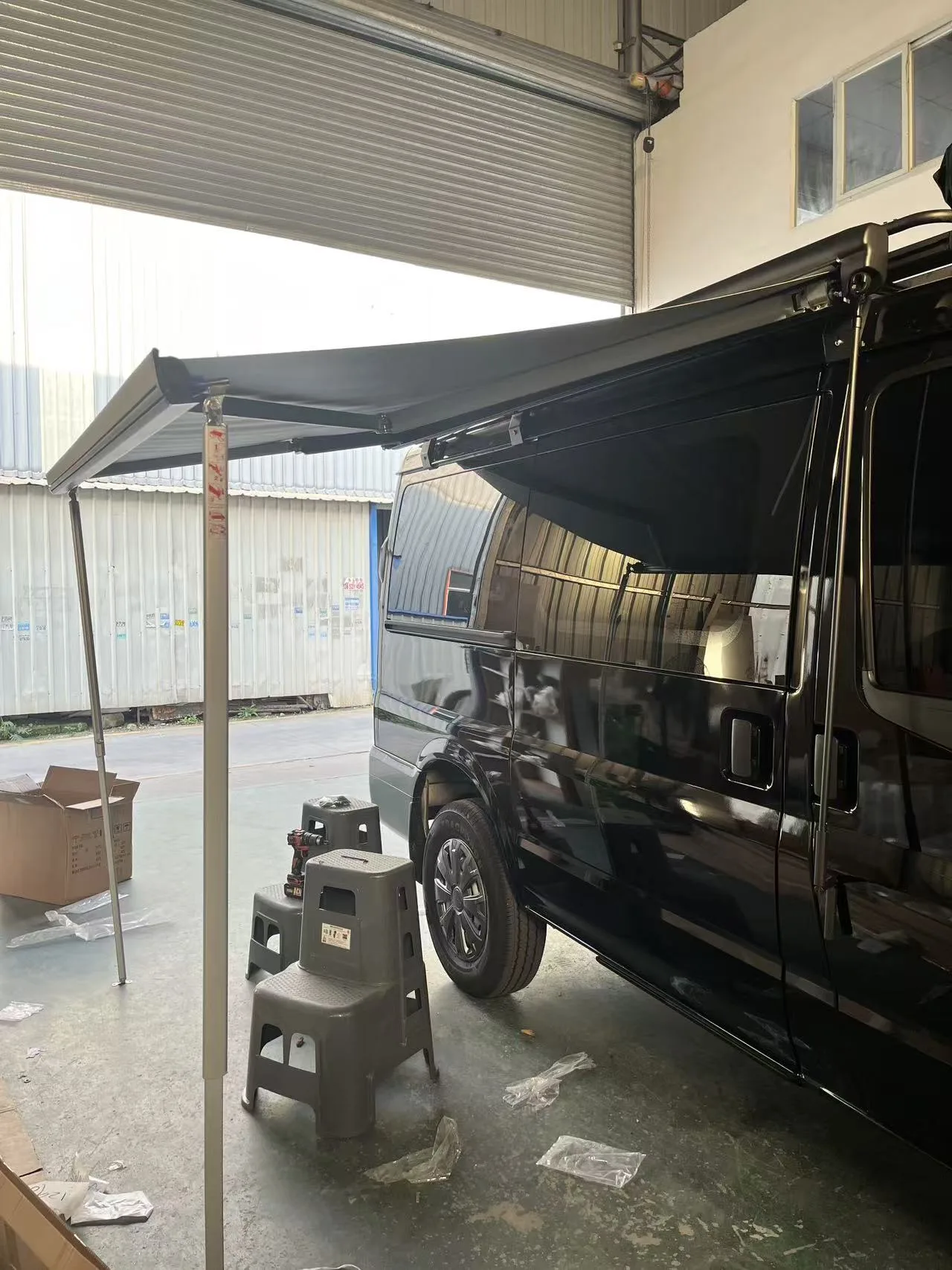 Black side awning extended beside an unidentified dark vehicle inside a workshop